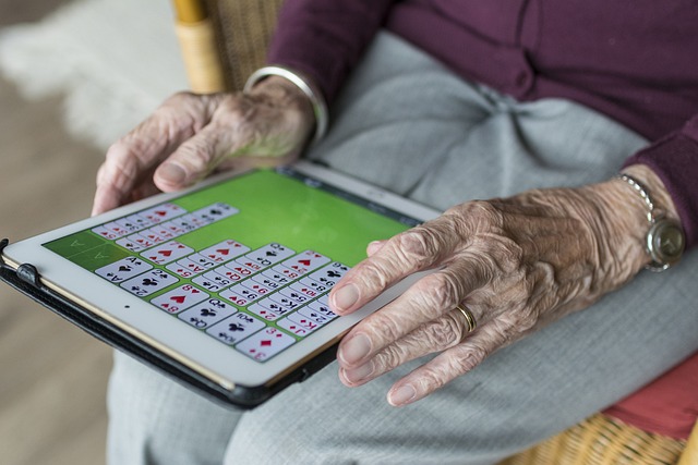 L'image montre une personne âgée (seules ses mains sont visibles), tenant une tablette numérique sur ses genoux et jouant à un jeu de cartes en ligne. 